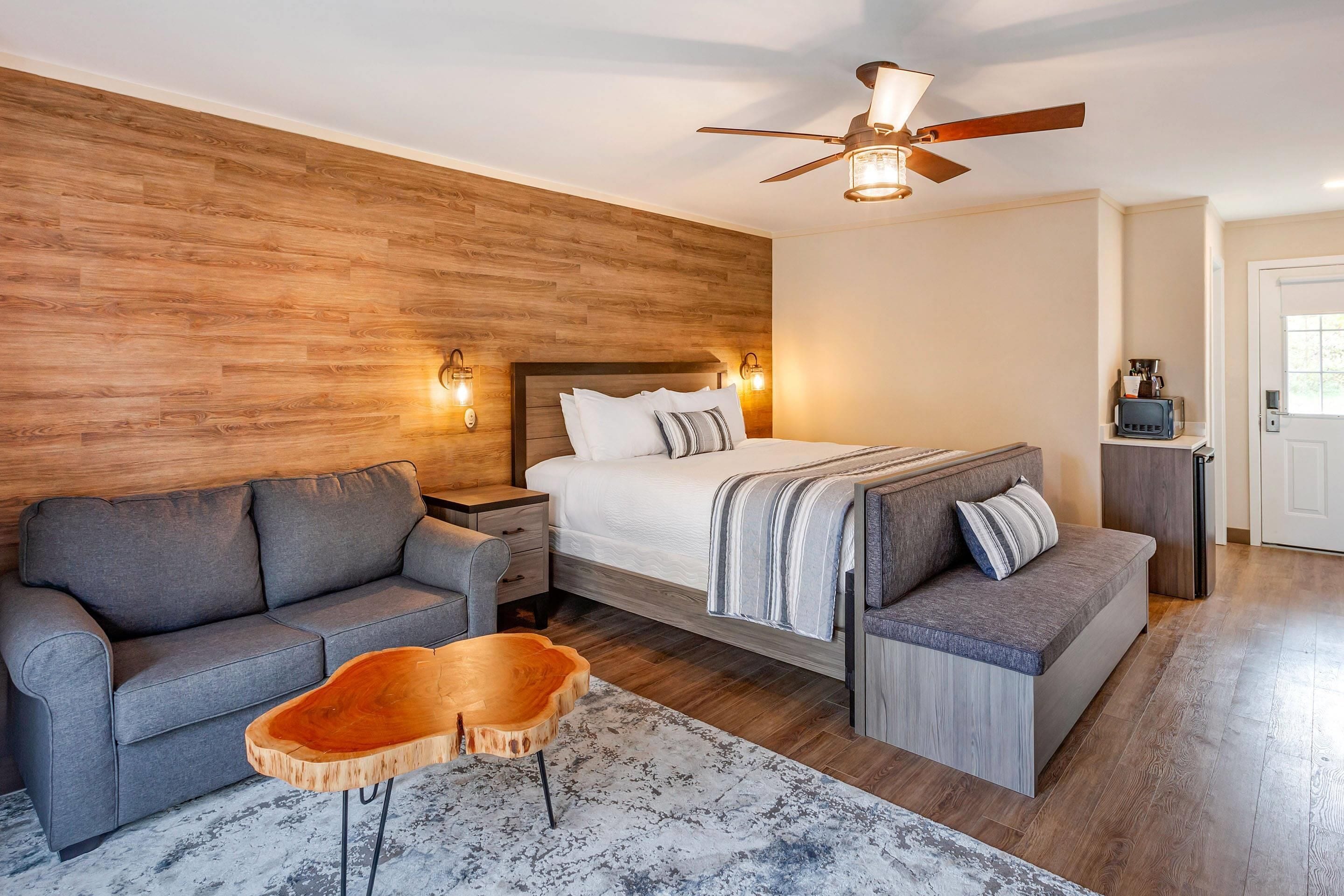 Un dormitorio con cabecera de madera, una cama, un sofá gris, una mesa de centro de madera y un ventilador de techo.