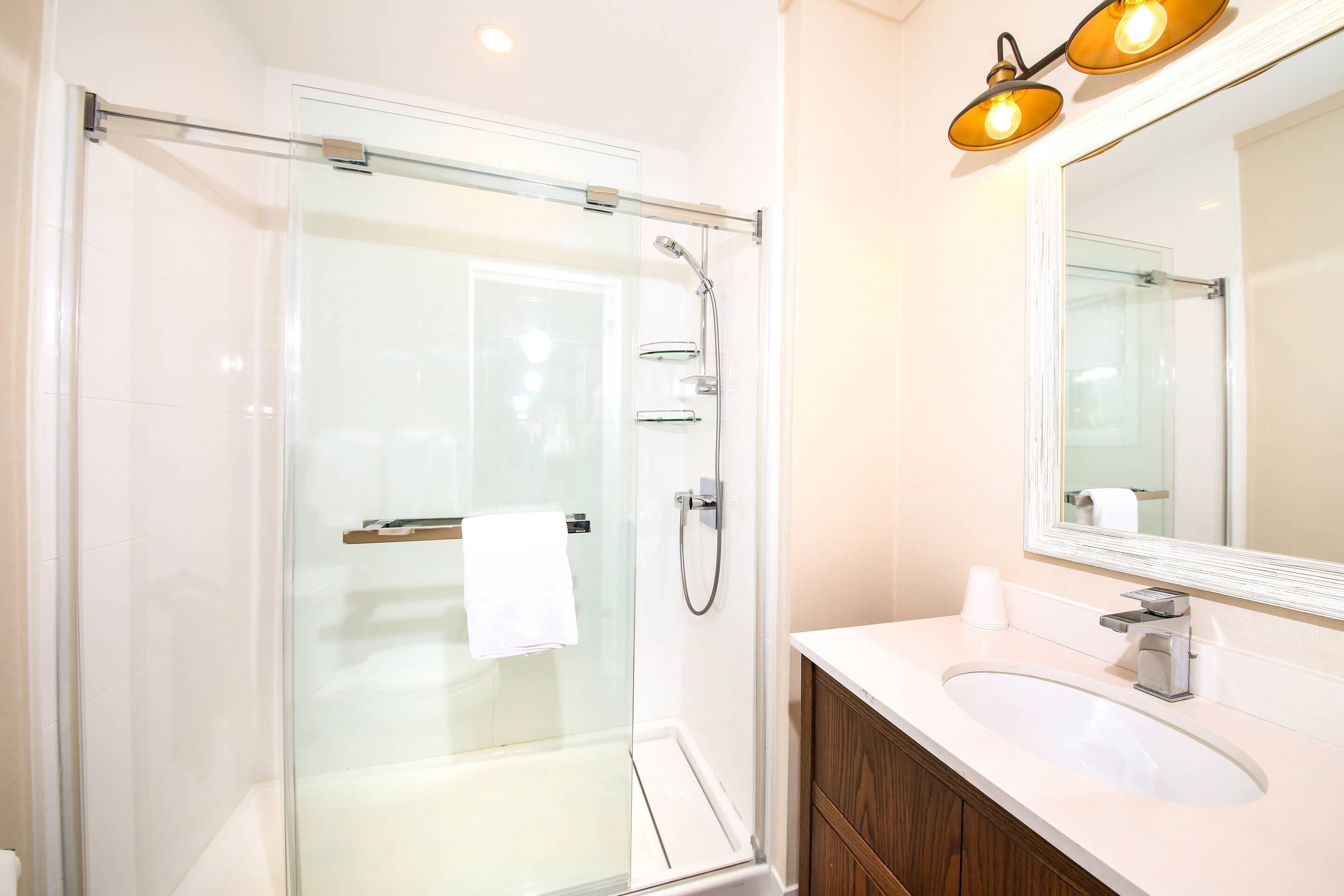 Un baño moderno con ducha envuelta en vidrio, un lavamanos blanco y un espejo con dos luces.