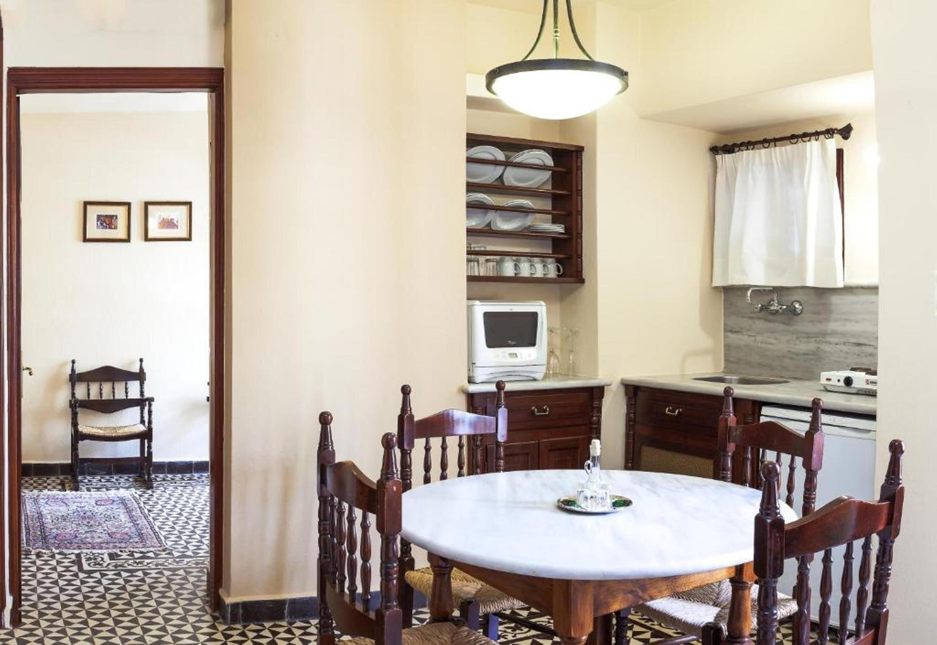 A compact kitchen with a round dining table, wooden chairs, a built-in shelf with dishes, a microwave, and a small refrigerator.