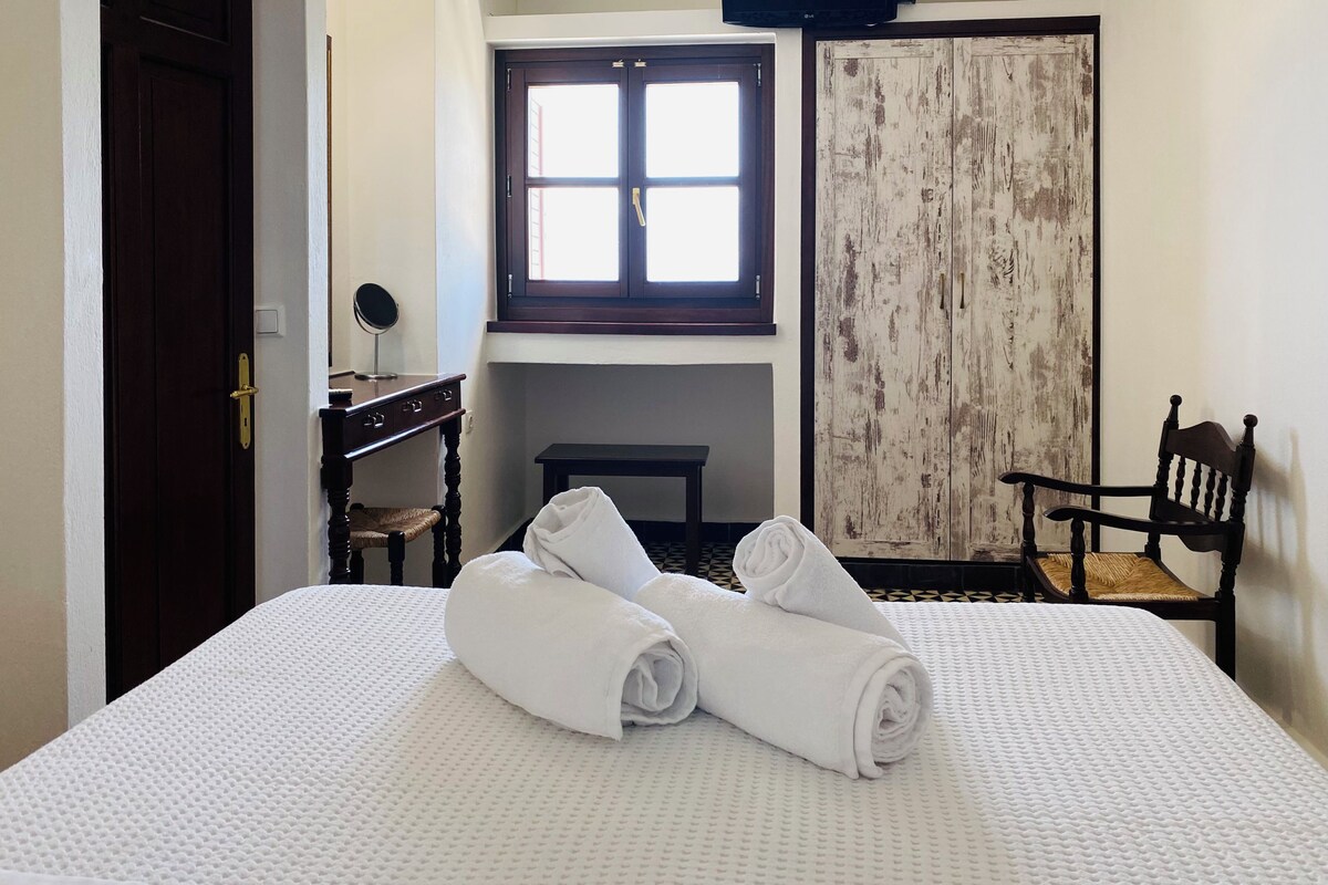 Une chambre d’hôtel avec un lit, une chaise en bois, une petite table avec une lampe, une armoire et une fenêtre.