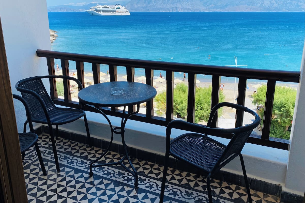 A balcony with two chairs and a table, overlooking a sea with a cruise ship in the distance and mountains in the background.