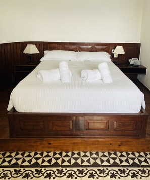Un lit bien fait, avec du linge de lit blanc et des serviettes pliées, une tête de lit en bois, des tables de chevet avec des lampes et un sol à motifs.