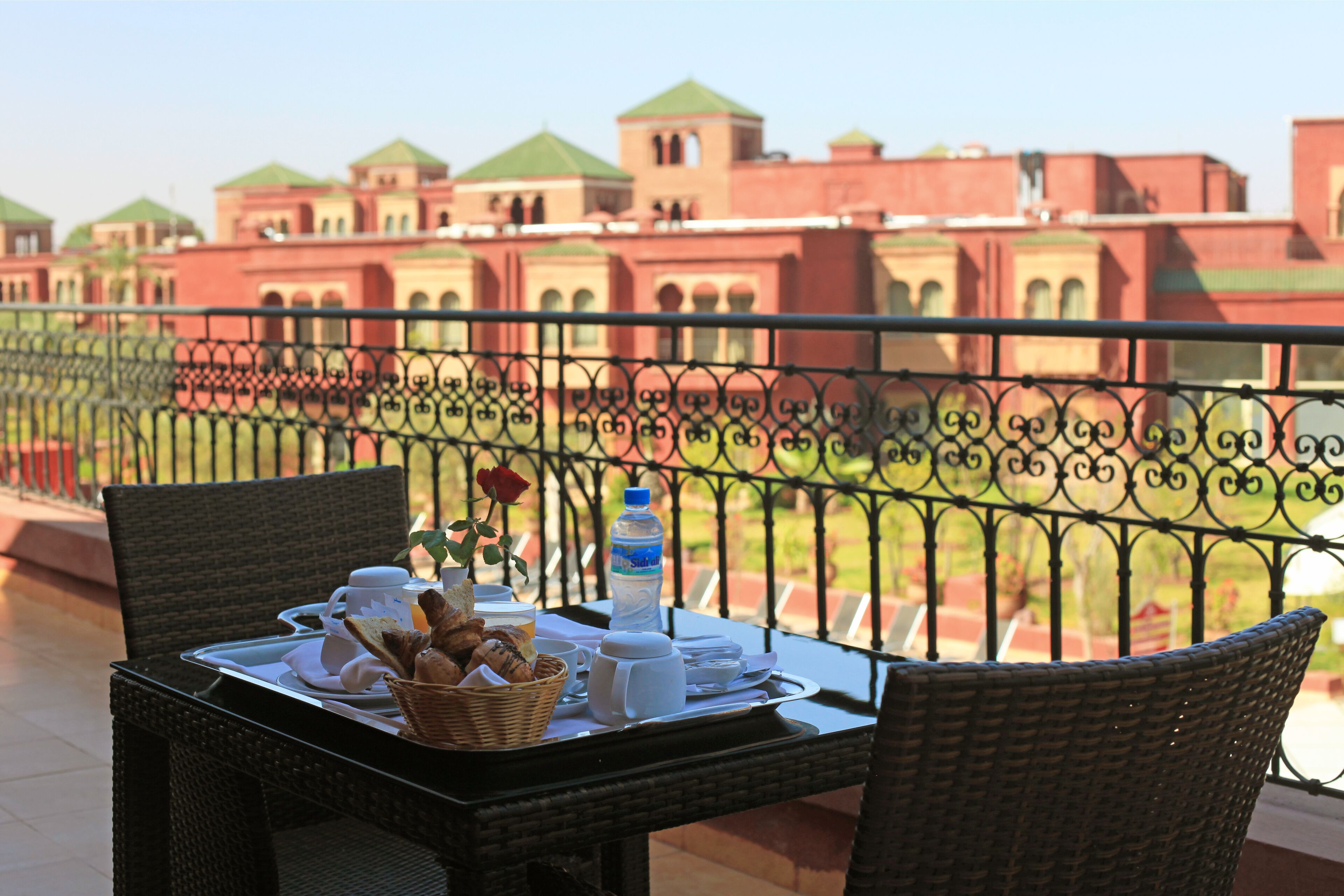 A dining table with breakfast items, a water bottle, and a basket of bread on a balcony with a view of a building with green roofs.