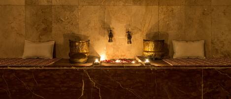 Un área de spa con paredes de mármol, dos cuencos dorados, velas y almohadones blancos.
