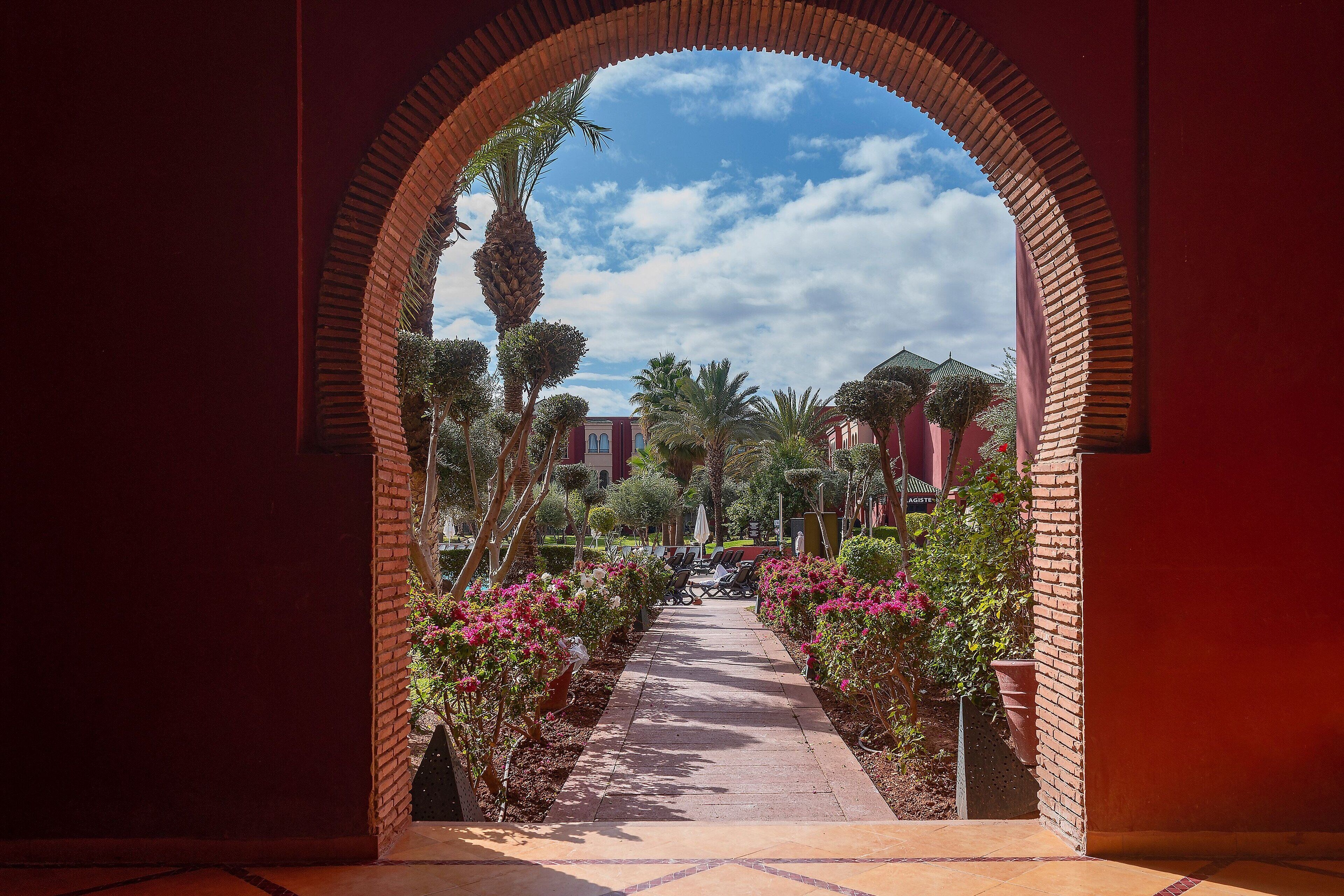 A pathway leading through a brick archway with a view of a courtyard, palm trees, and a building in the background.