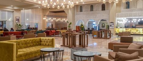 A spacious lobby with a yellow sofa, multiple tables, and a high ceiling with hanging lights.