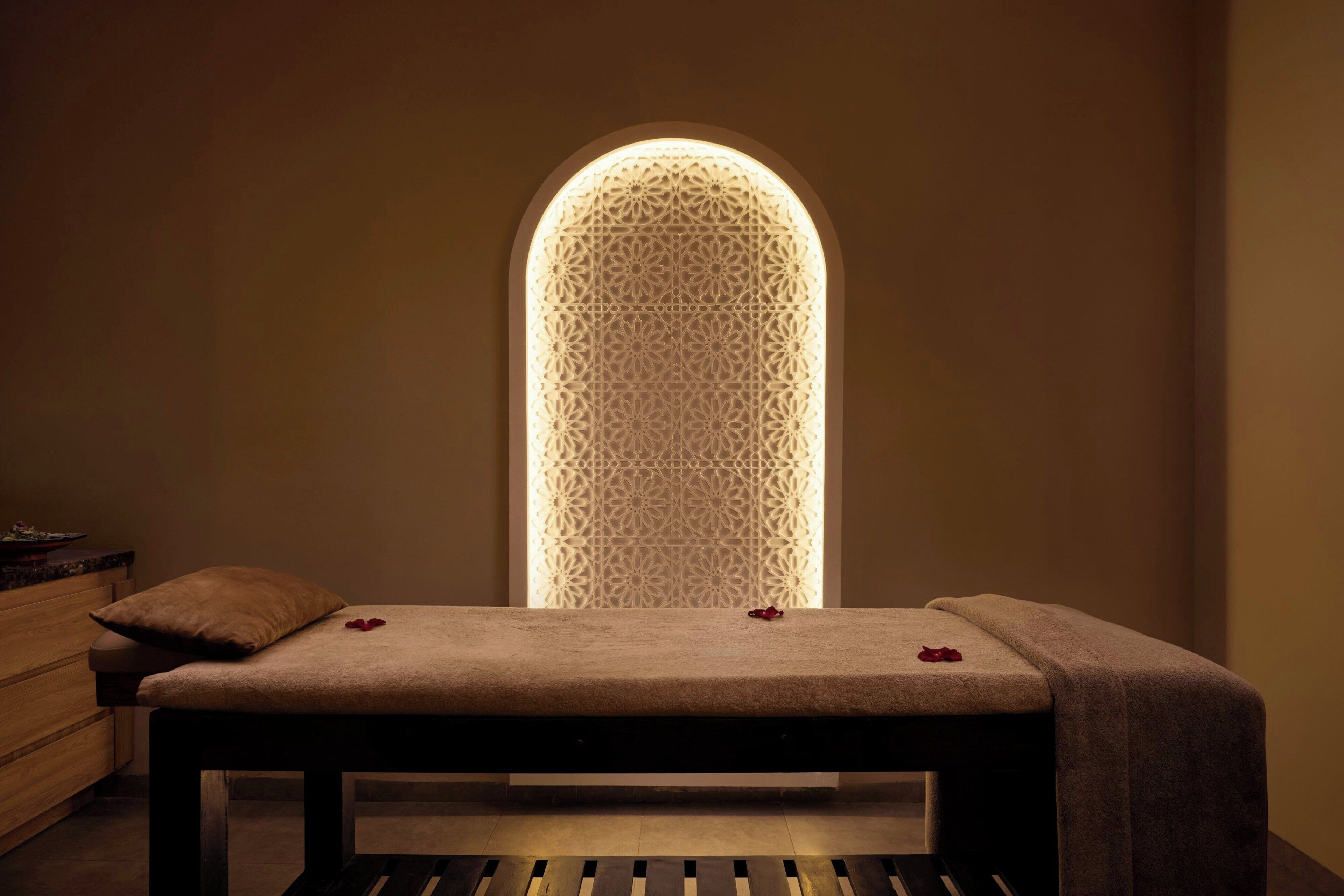 A room with a bed, a wooden bench, and a wall with a geometric light pattern.