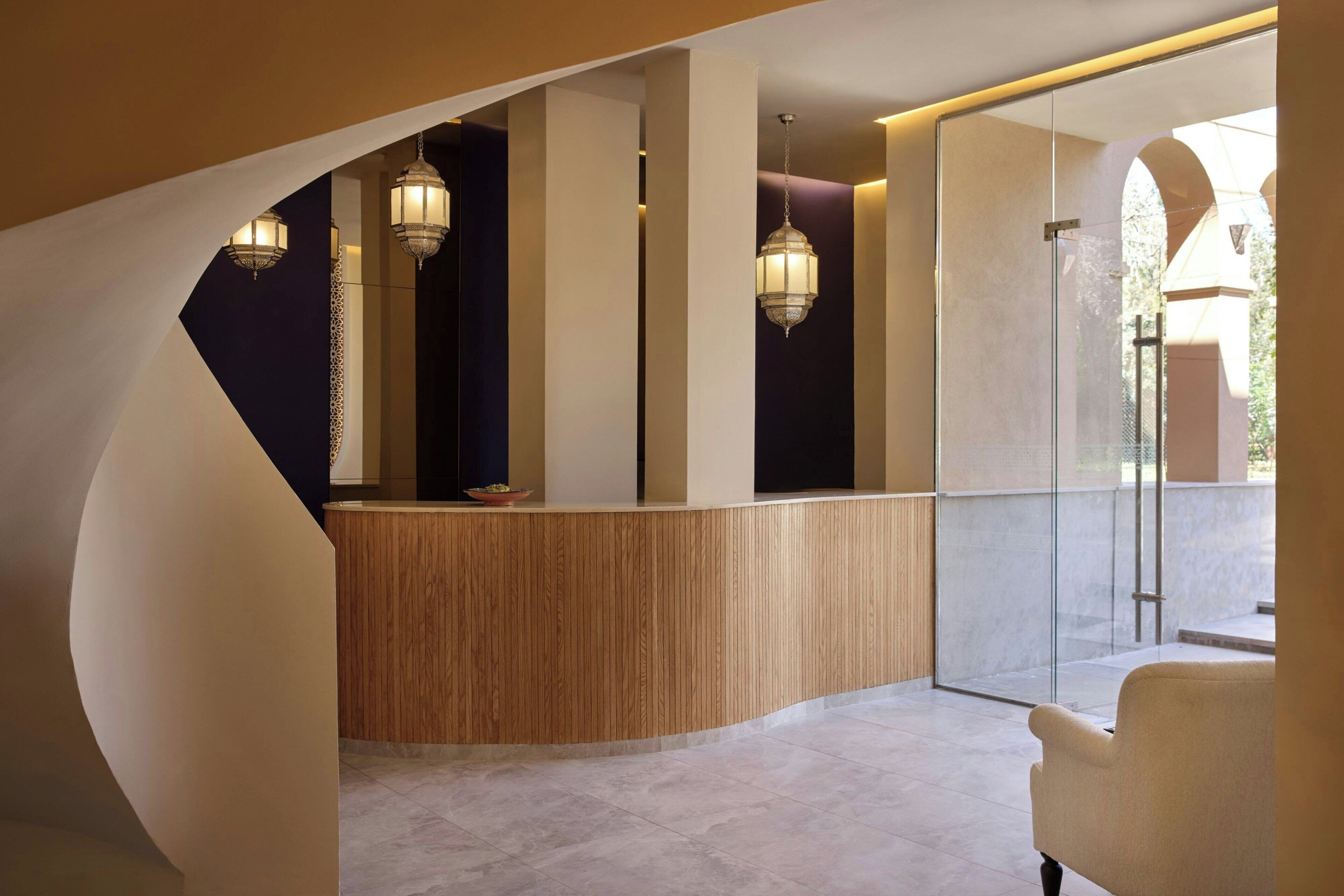 Zona de recepción con mostrador curvo de madera, faroles colgantes y puerta de cristal que da a un espacio exterior.