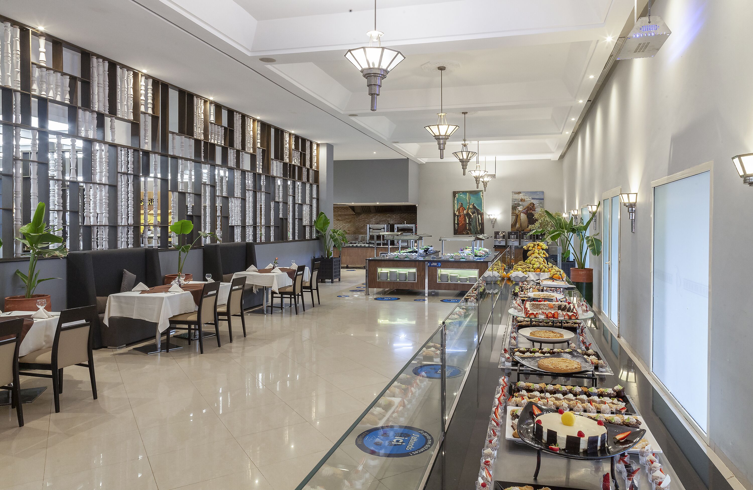 A modern restaurant with a long counter displaying an assortment of desserts, a seating area with tables and chairs, and a decorative wall with patterned shelves.