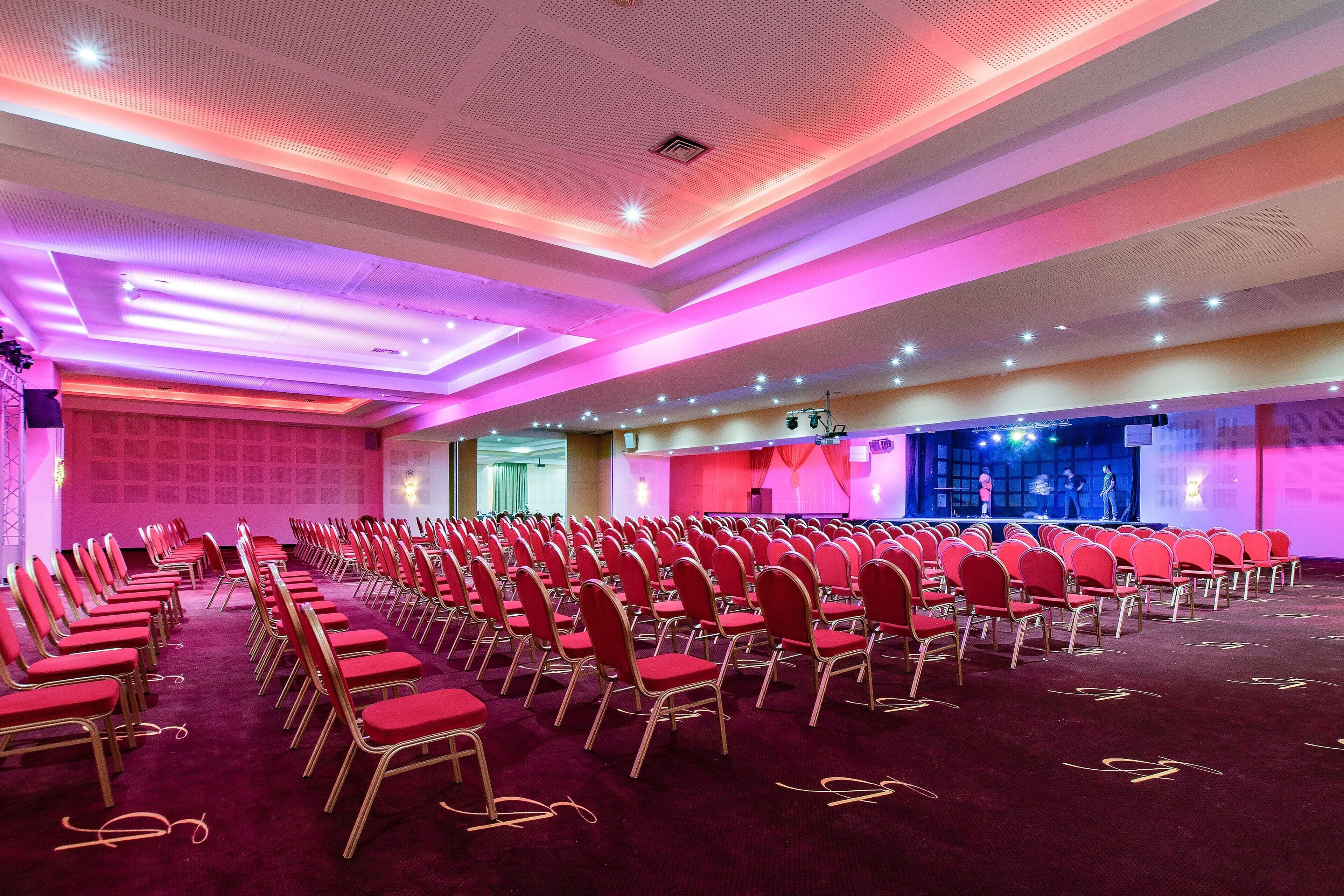 Un salón de conferencias con filas de sillas rojas y un escenario con iluminación.
