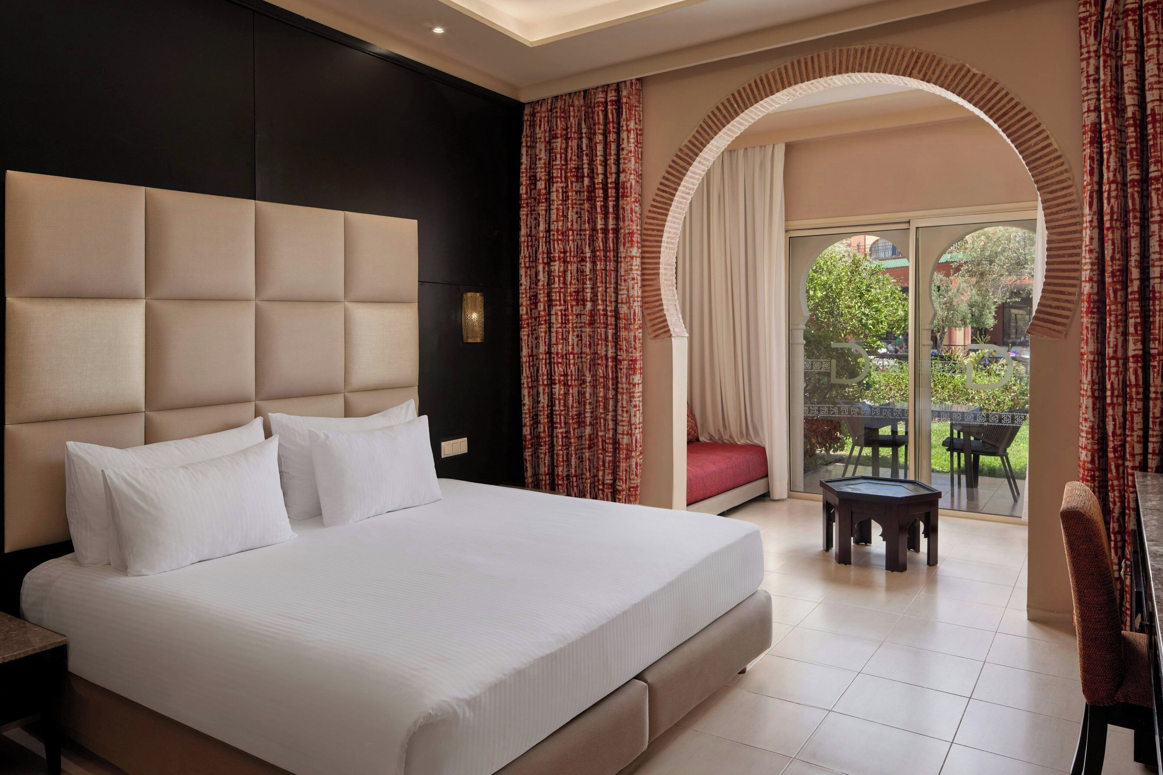 A hotel room with a large bed, a desk, a chair, a small table, and a view of the outdoors through an arched doorway.