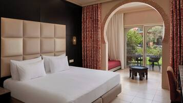 Quarto de hotel com uma cama grande, uma escrivaninha, uma cadeira, uma mesinha e vista para o exterior através de uma porta arqueada.