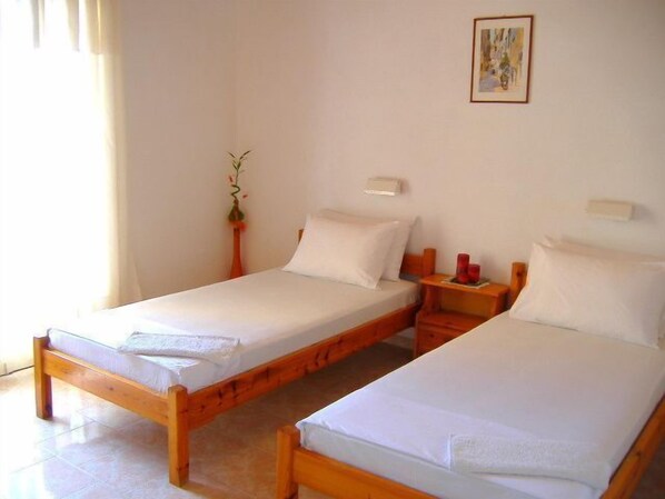 A room with two single beds, wooden headboards, white linens, a small bedside table with a red candle, and a wall-mounted light fixture.