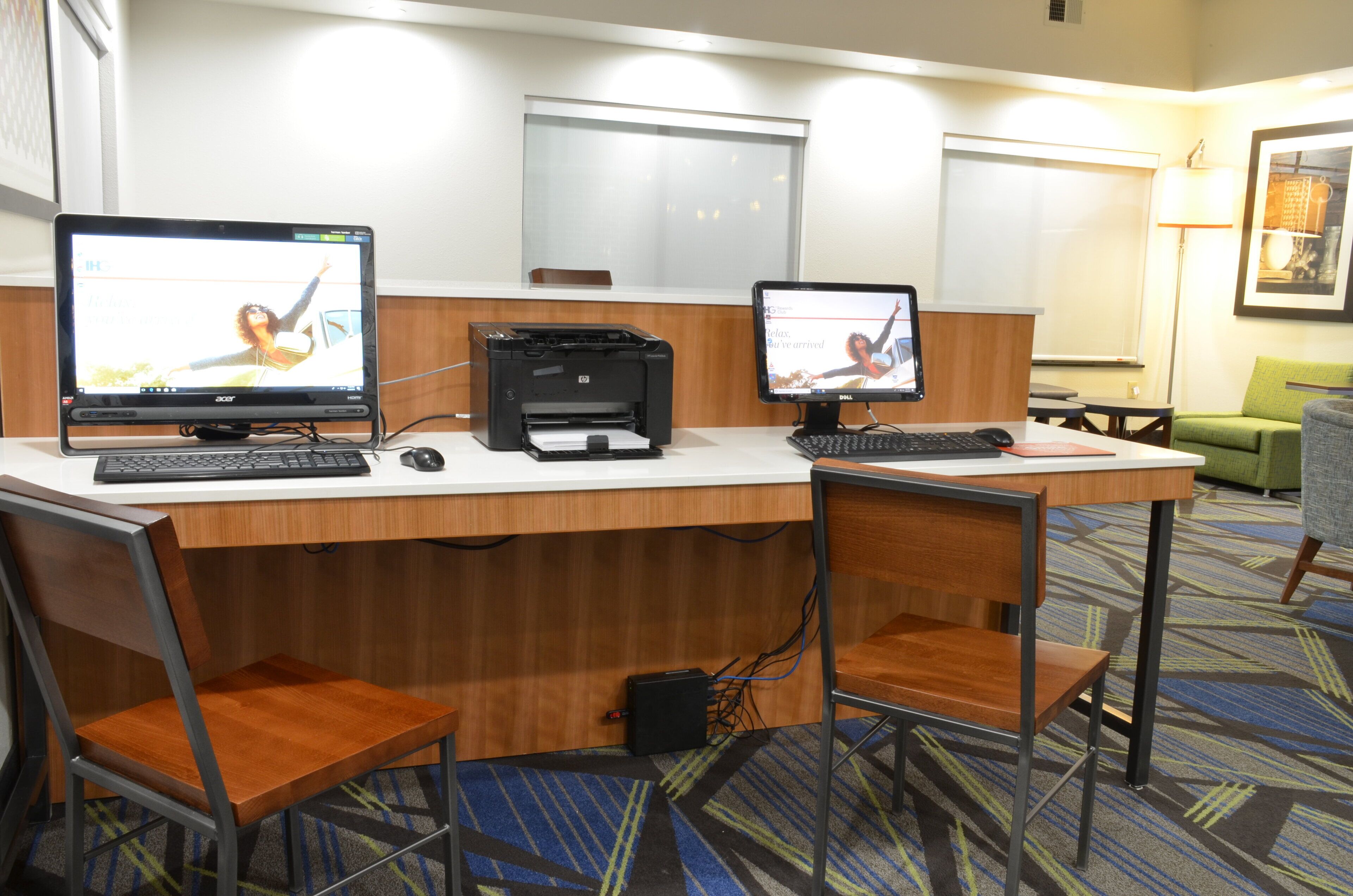 A hotel workspace with two computers, a printer, and a desk with chairs.