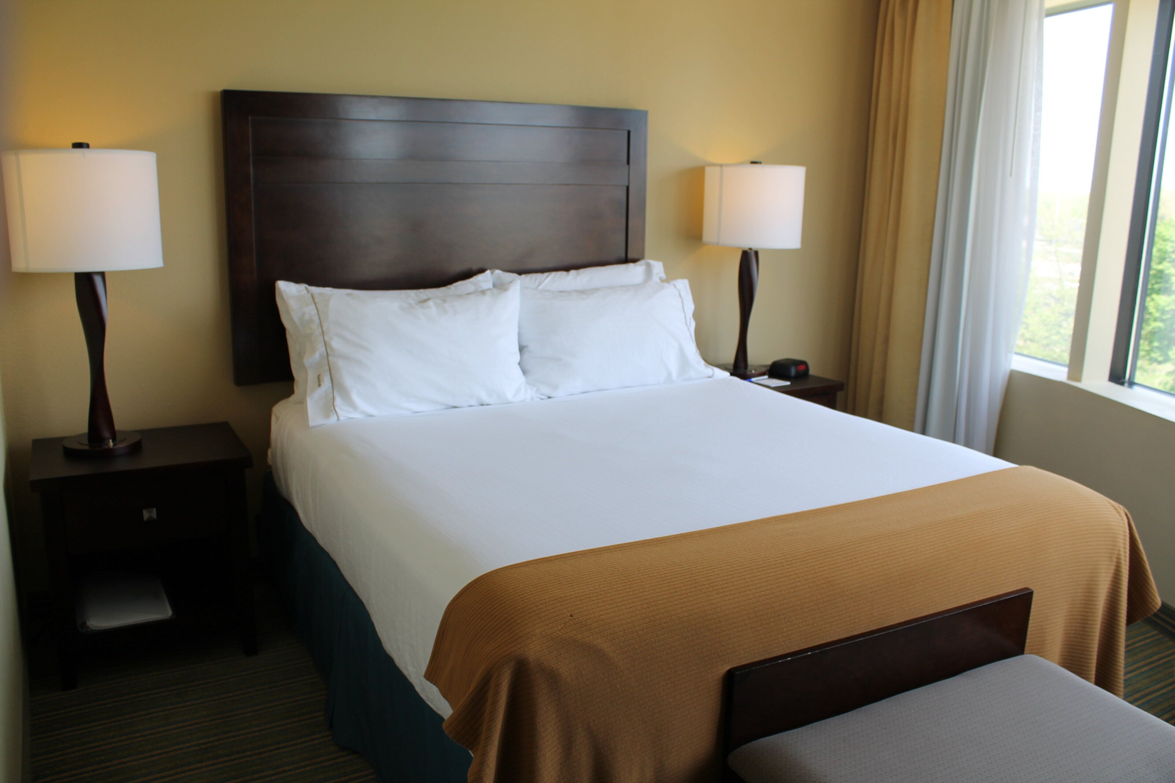 A hotel room with a large bed, two bedside tables, a nightstand, a window with curtains, and a view of greenery outside.