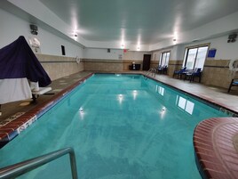 An indoor swimming pool with a tiled floor, a lifeguard chair, and a poolside area with chairs and a notice board.