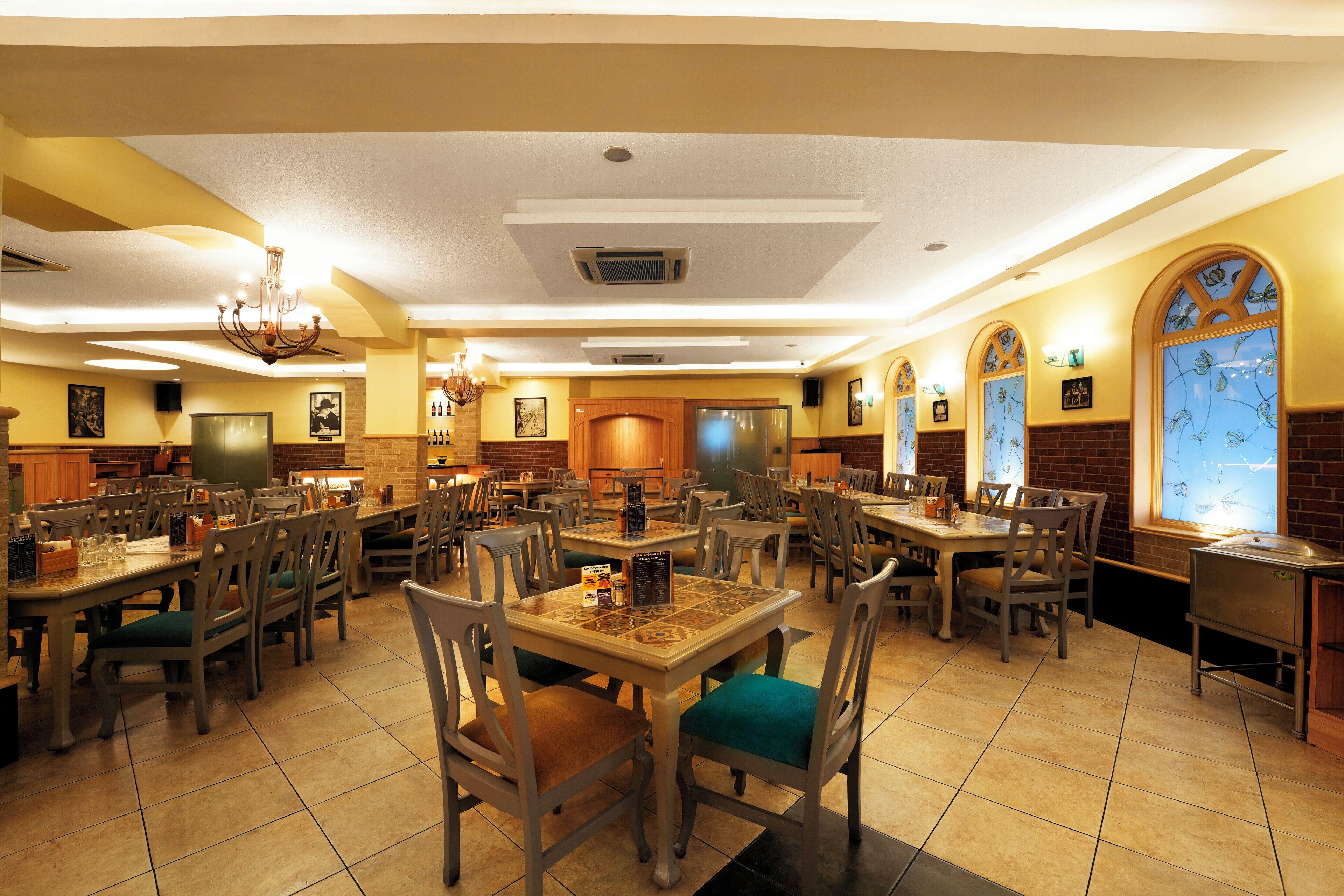 A spacious restaurant with multiple dining tables and chairs, a chandelier, and decorative windows.