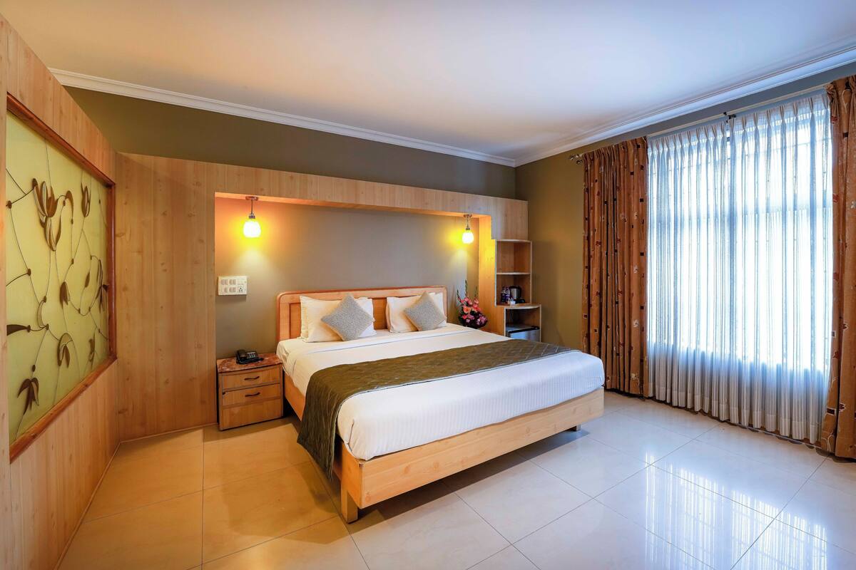 A hotel room with a wooden bed, a nightstand, a shelf with toiletries, and a large window with curtains.