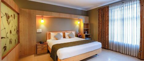A hotel room with a wooden bed, a nightstand, a shelf with toiletries, and a large window with curtains.