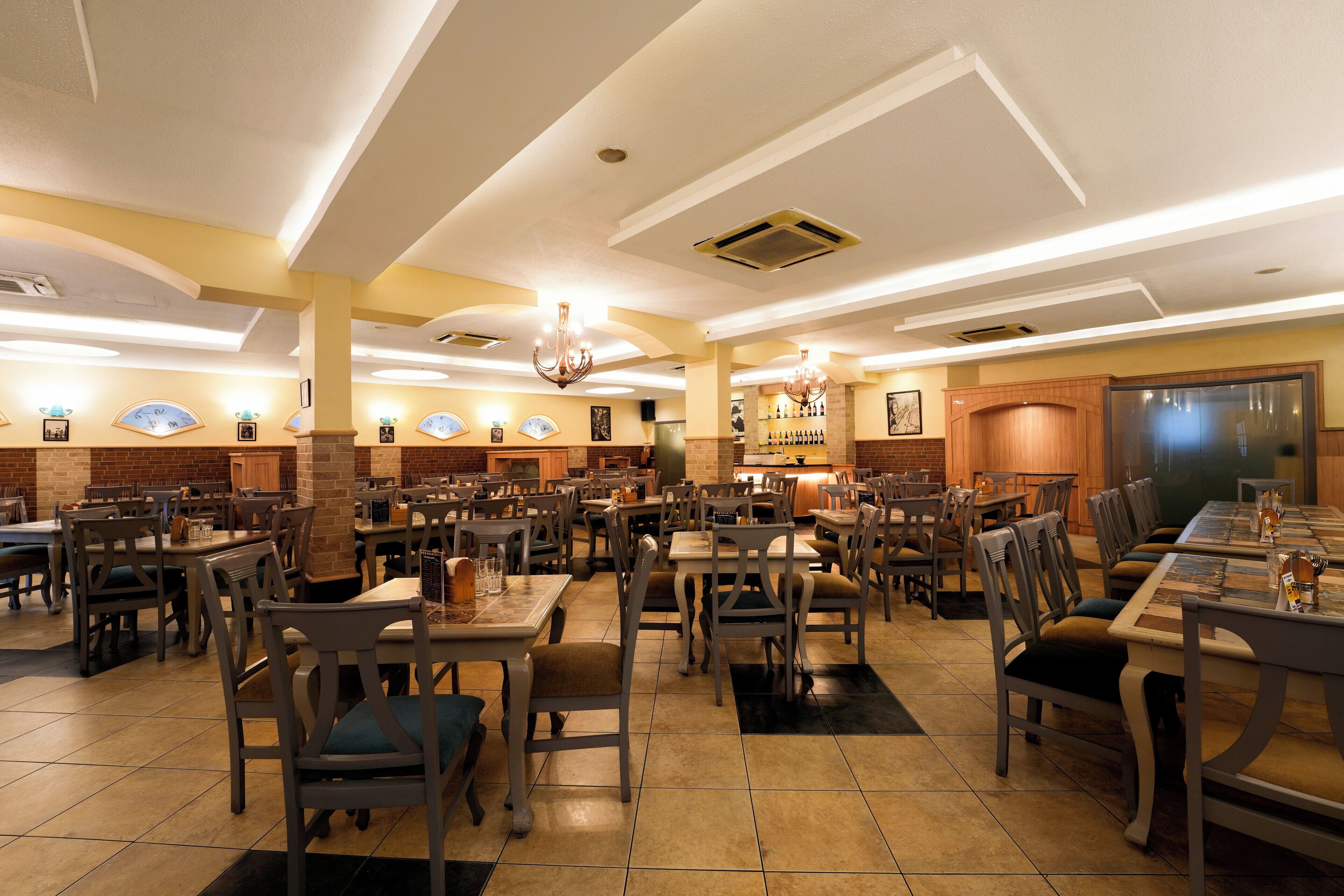A spacious restaurant with multiple dining tables and chairs, a bar area, and a kitchen in the background.