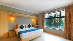 A bedroom with a large bed, wooden headboard, bedside tables, a nightstand, a window with curtains, and a view of greenery outside. - Cross Roads Inn (Bengaluru)
