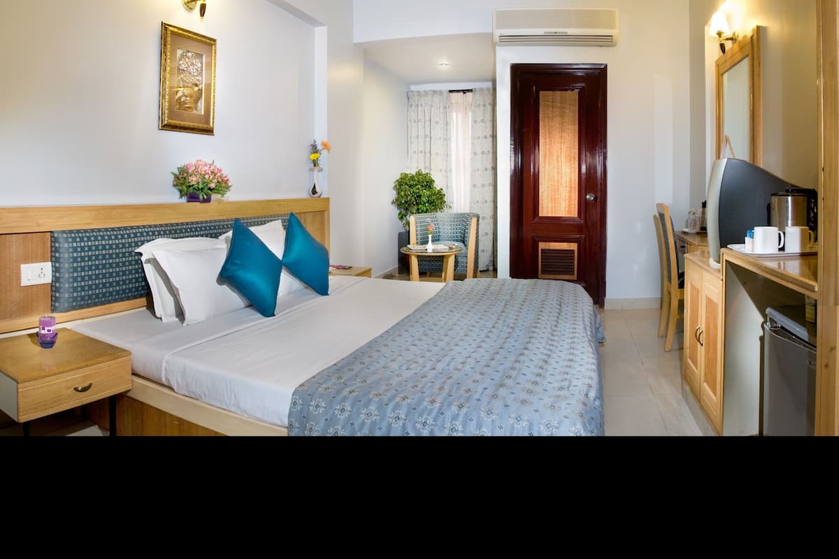 A hotel room with a bed, bedside table, chair, small table with flowers, and a desk with a kettle.