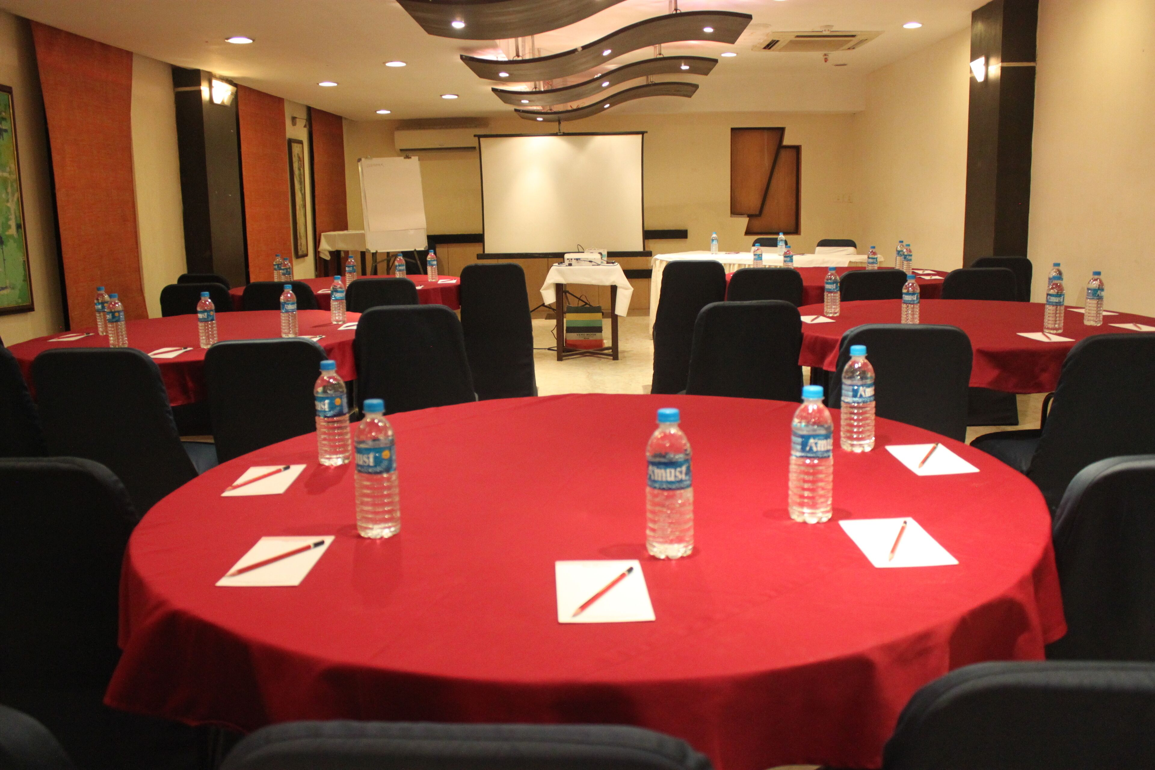 Un salón de conferencias con varias mesas cubiertas con manteles rojos, cada una con botellas de agua y blocs de notas.