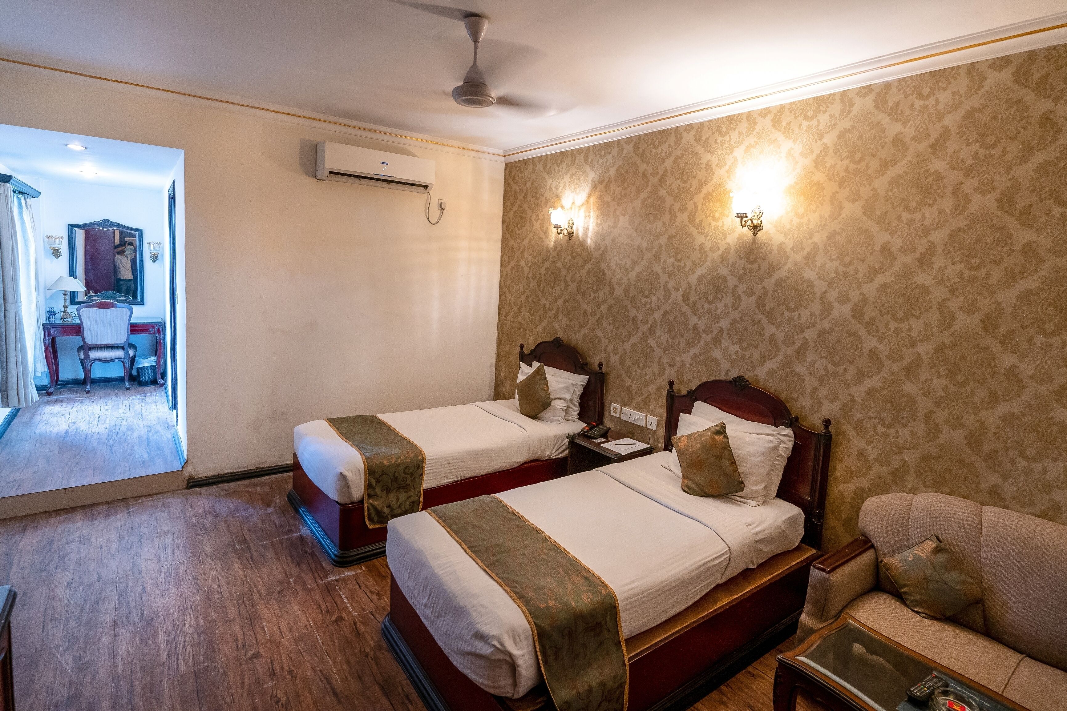 A hotel room with two single beds, a wooden floor, a sofa, a small table, and a mirror.