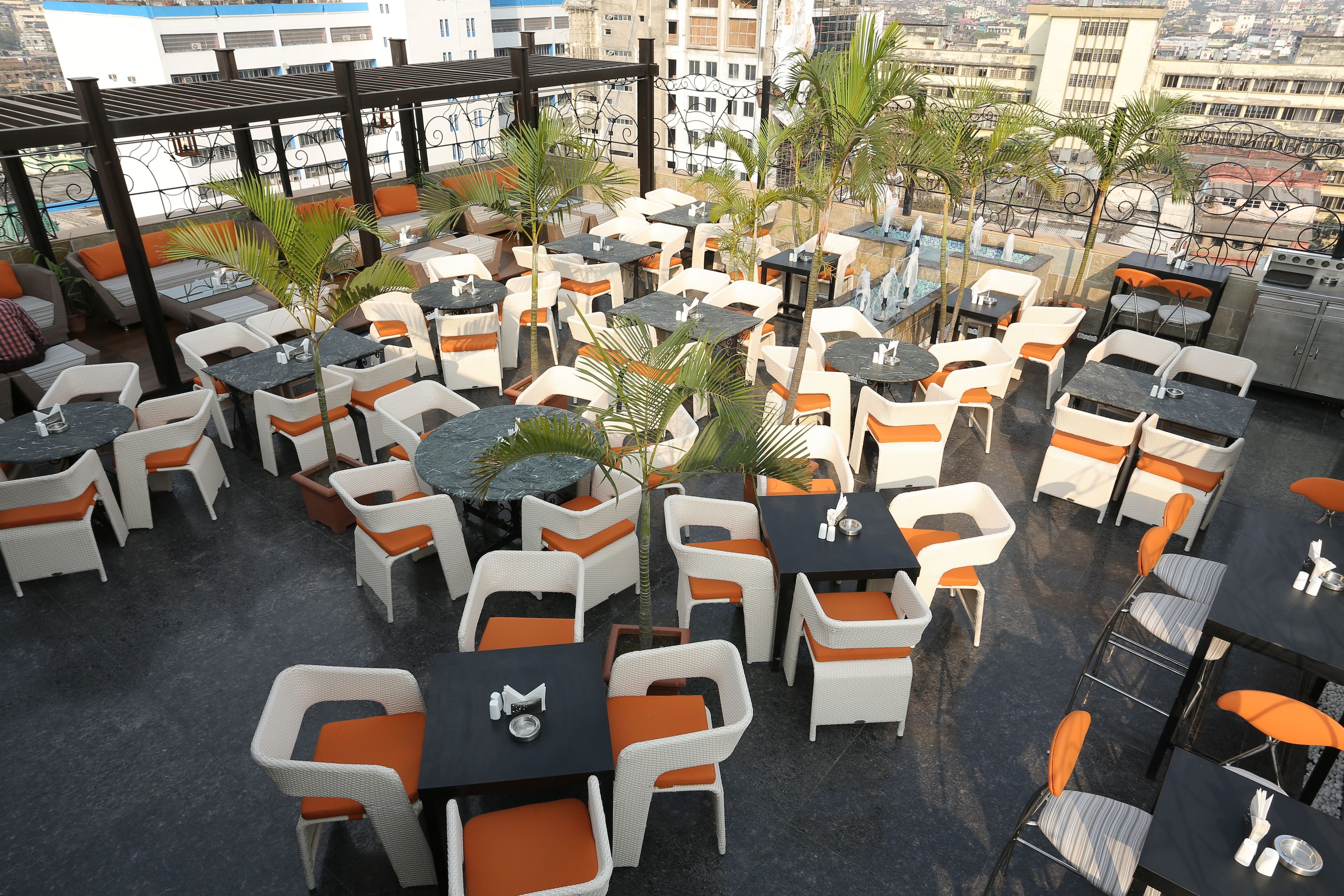 Une terrasse sur le toit avec des espaces de détente en plein air, comprenant des chaises et des tables blanches et orange, entourée de palmiers et offrant une vue sur la ville.