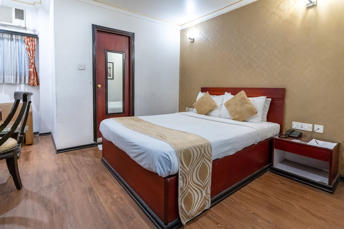 Ein Hotelzimmer mit Holzboden, einem Bett mit weißer Bettwäsche und braunen Kissen, einem Nachttisch mit Telefon und Unterlagen sowie einer Tür mit Spiegel.