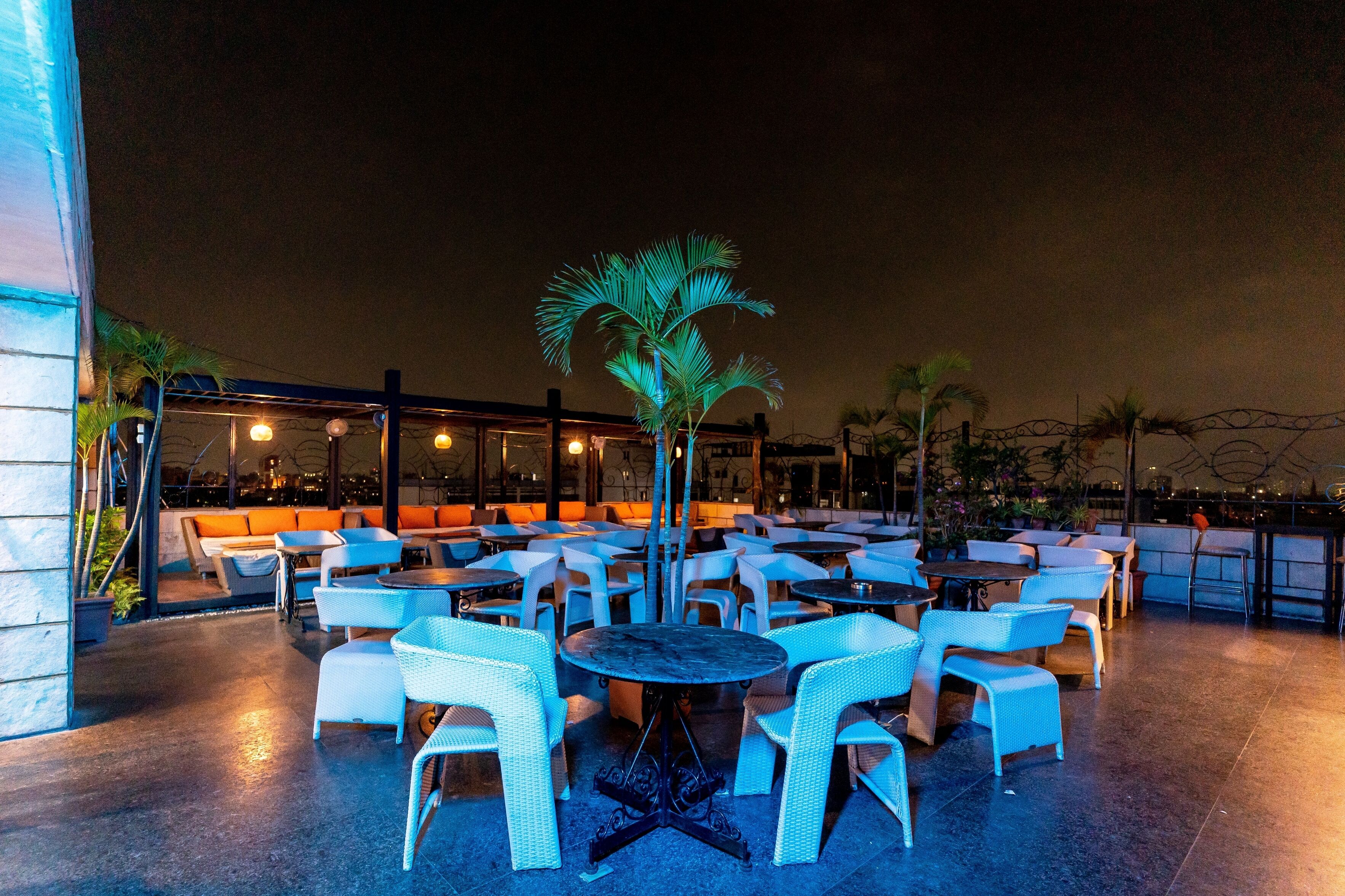 Un espace extérieur aménagé avec des tables rondes et des chaises en osier blanc, entouré de palmiers et offrant une vue sur la ville la nuit.