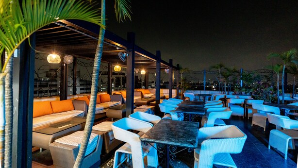 An outdoor seating area with tables and chairs, covered by a roof, and surrounded by palm trees. - The Lindsay (Kolkata)