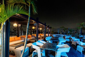 An outdoor seating area with tables and chairs, covered by a roof, and surrounded by palm trees. - The Lindsay (Kolkata)