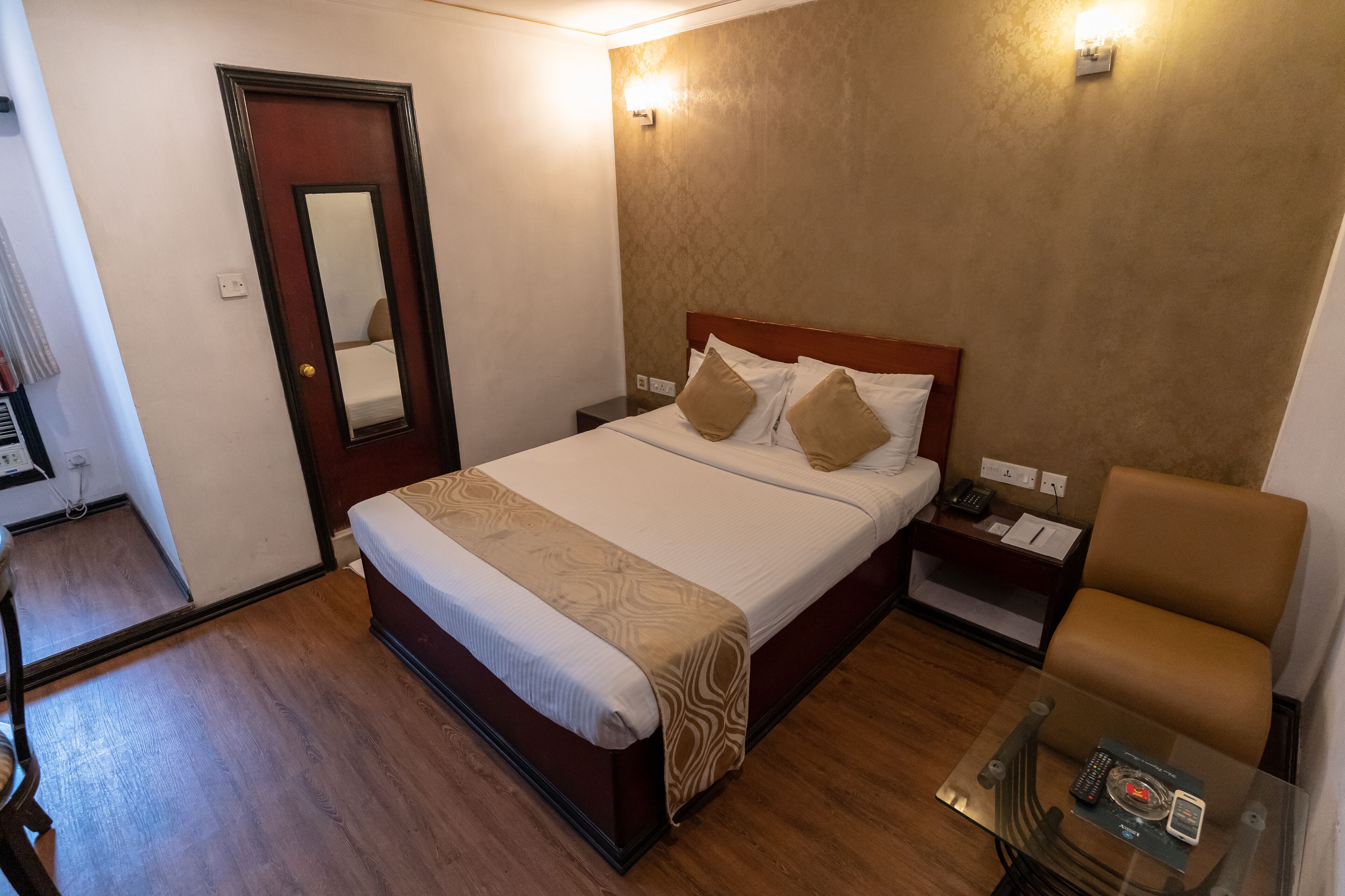 A hotel room with a wooden floor, a bed with a patterned bedspread, a bedside table with a lamp, and a chair.