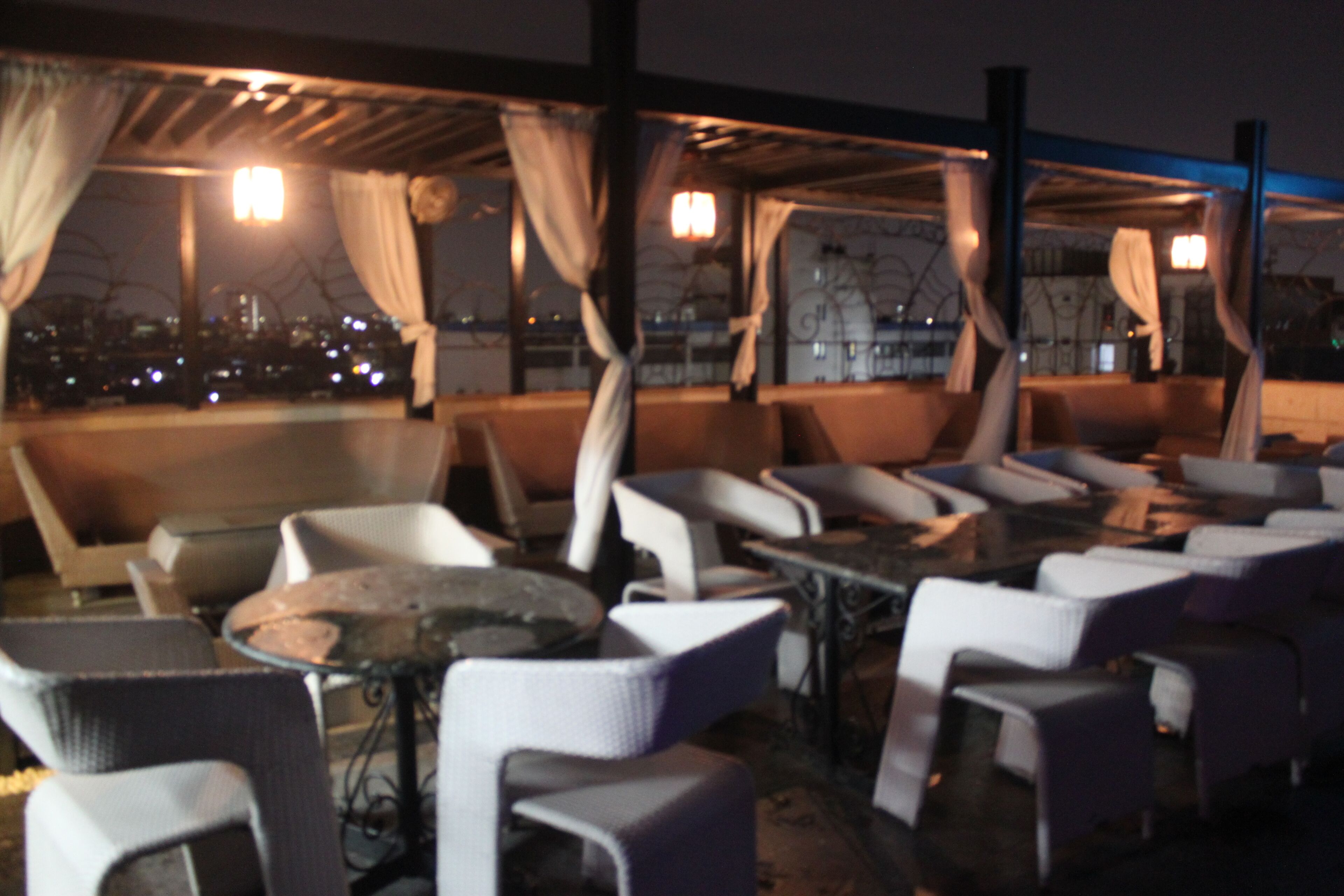 Un espacio al aire libre con sillas y mesas blancas, cubierto por una pérgola con cortinas, con vista a un paisaje urbano nocturno.