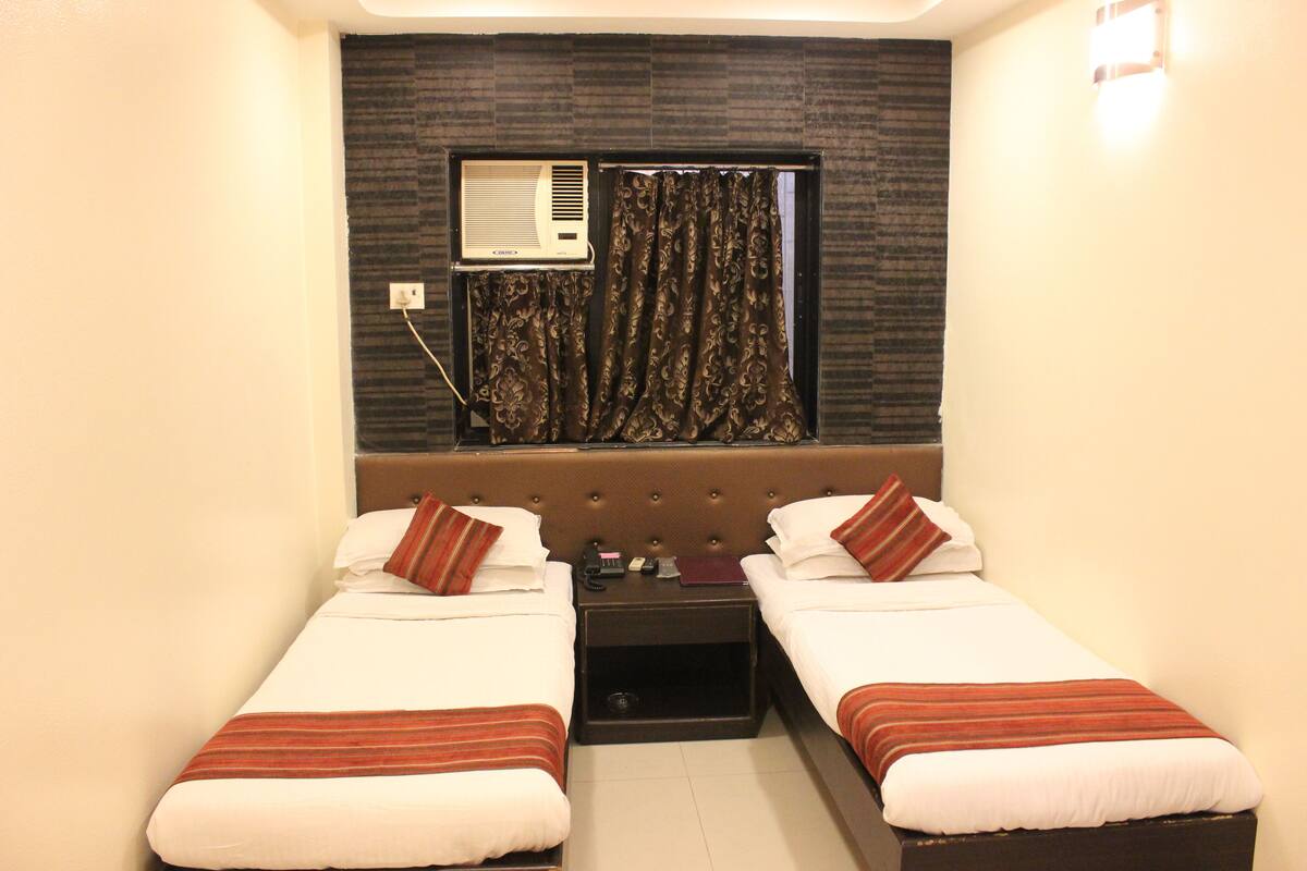 Habitación de hotel con dos camas individuales, cabecera oscura, unidad de aire acondicionado, mesita de noche y ventana con cortinas estampadas.
