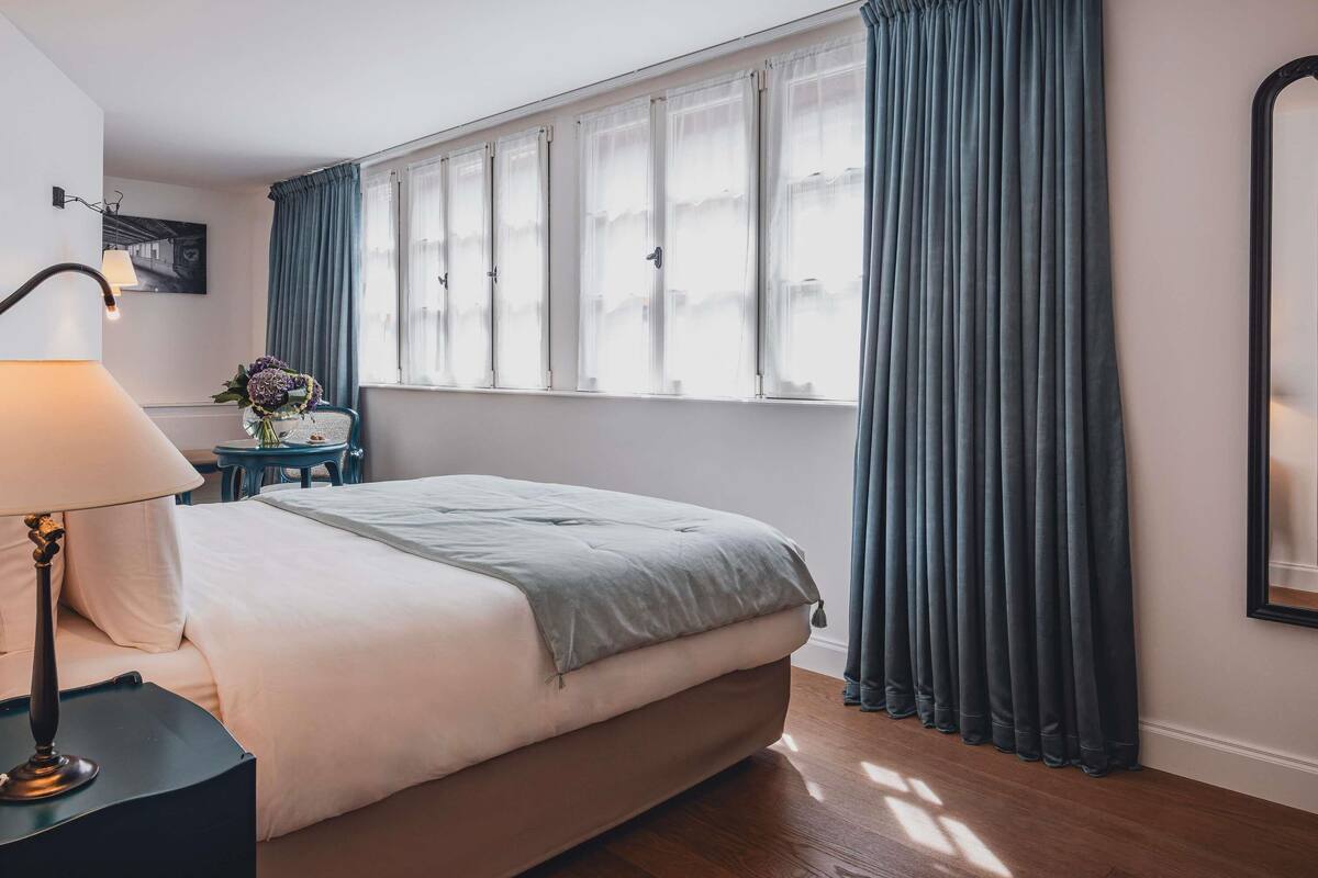 A neatly arranged bedroom with a bed, a bedside table with a lamp, a small table with a vase of flowers, and a window with curtains.