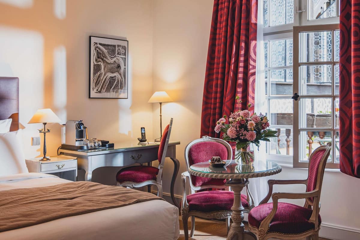 A hotel room with a bed, two chairs, a small table with a vase, a desk with a lamp, and a window with curtains.