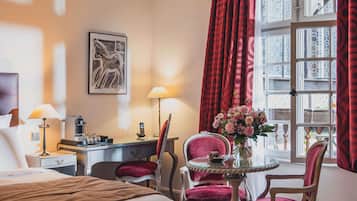 A hotel room with a bed, two chairs, a small table with a vase, a desk with a lamp, and a window with curtains.
