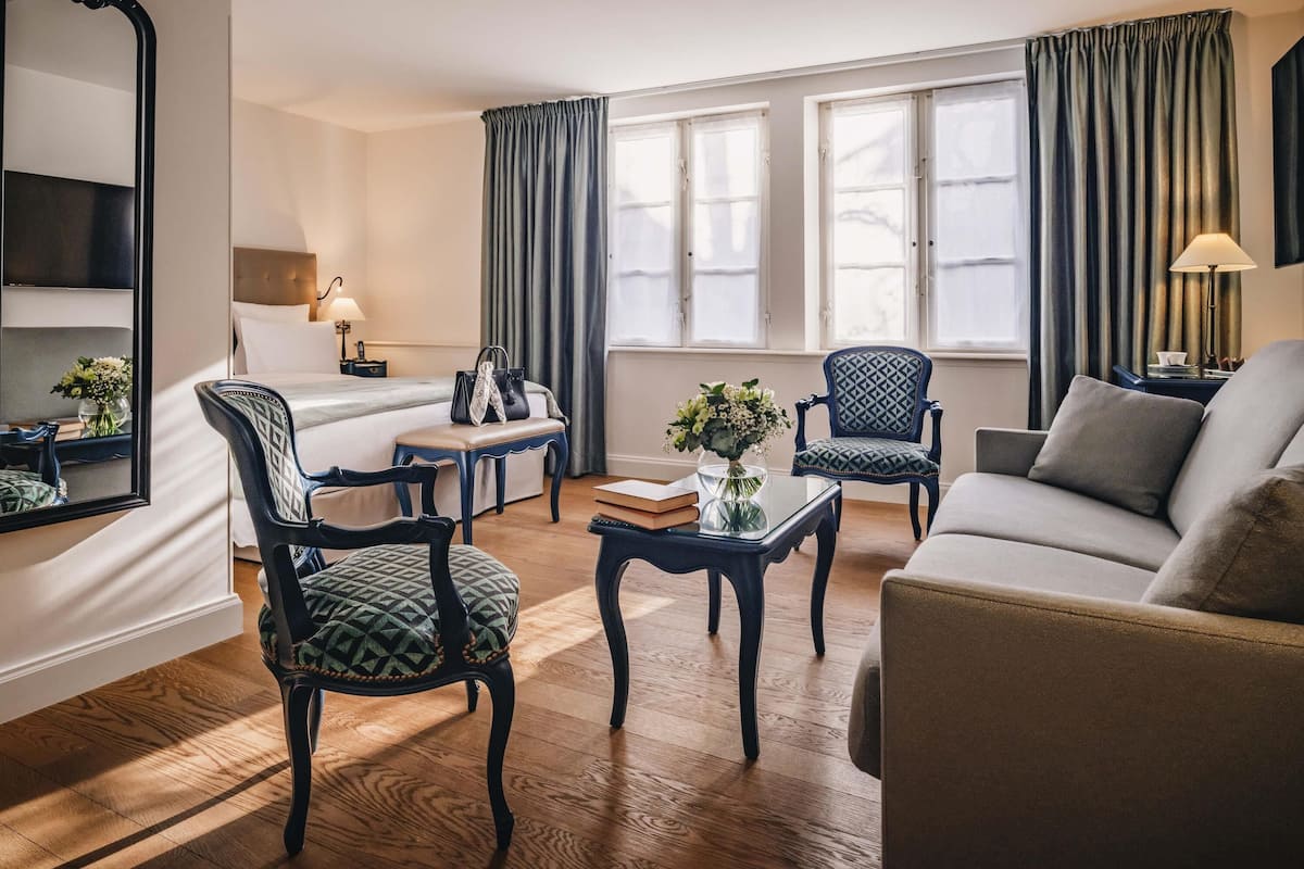A hotel room with a bed, two armchairs, a sofa, a small table, a mirror, and a window with curtains.
