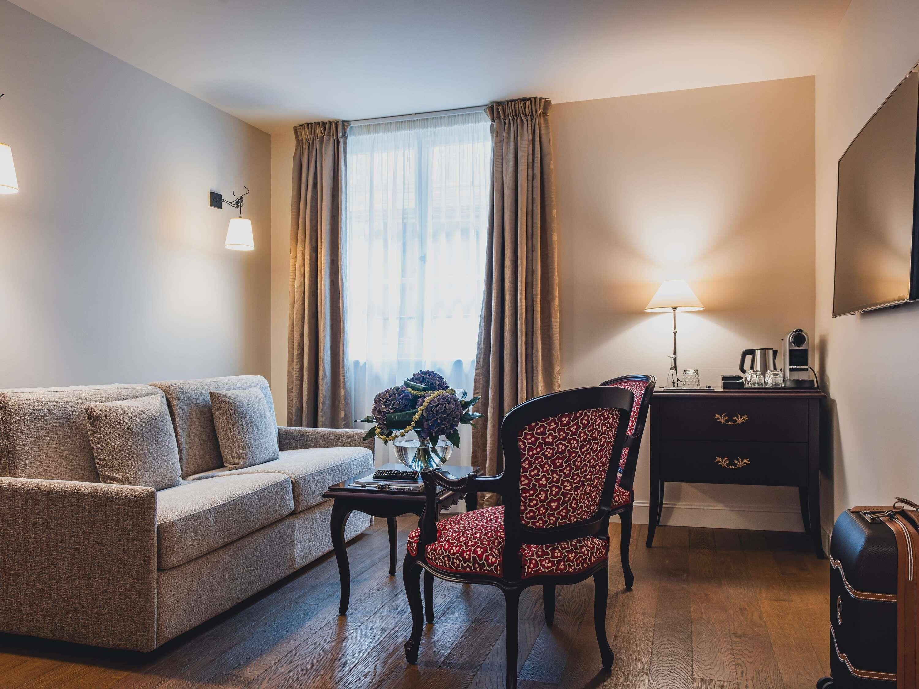 A furnished room with a sofa, a chair, a small table, a lamp, a desk, and a suitcase.