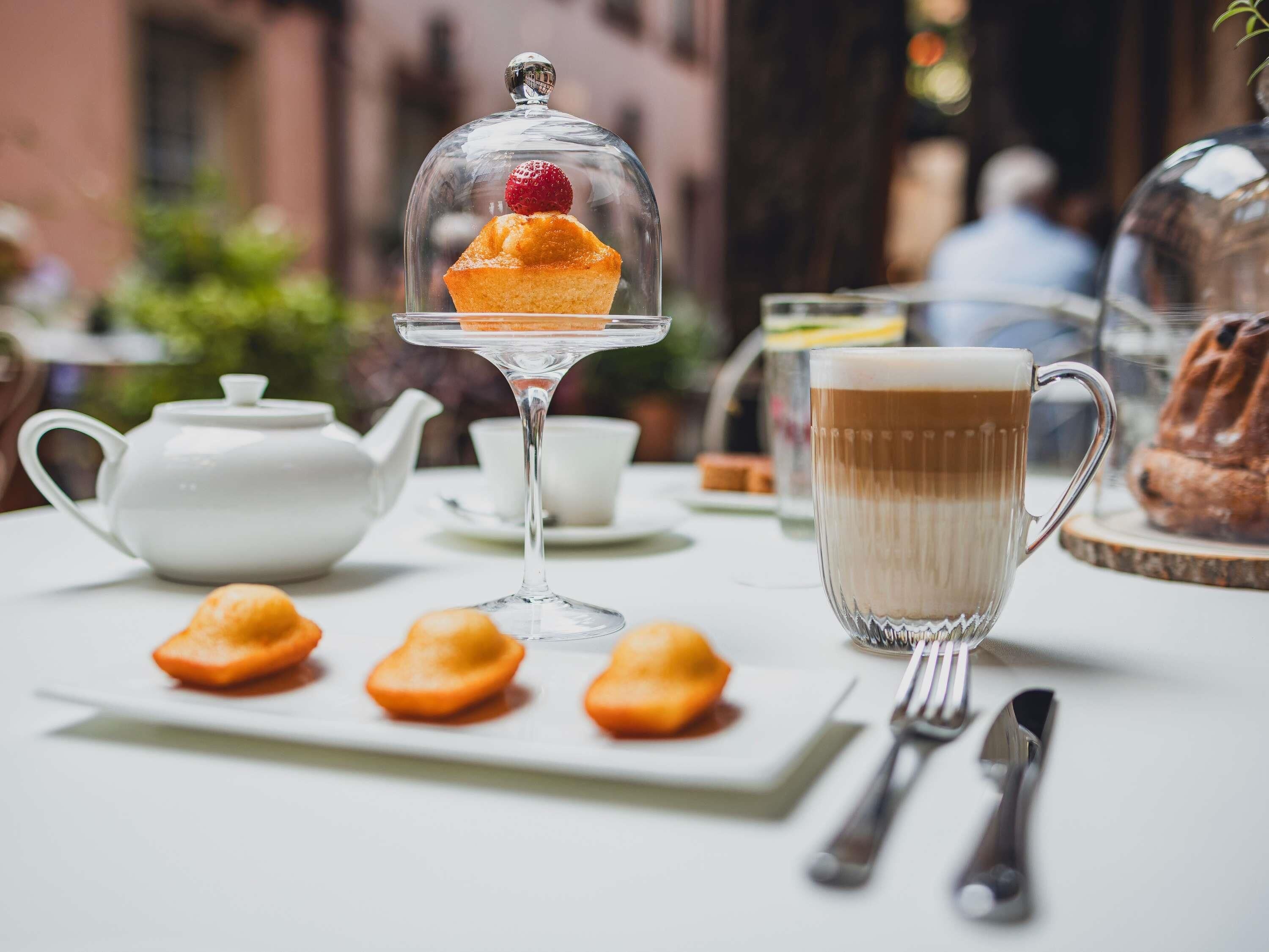 A table with a teapot, a cup of coffee, a dessert in a glass dome, and pastries.