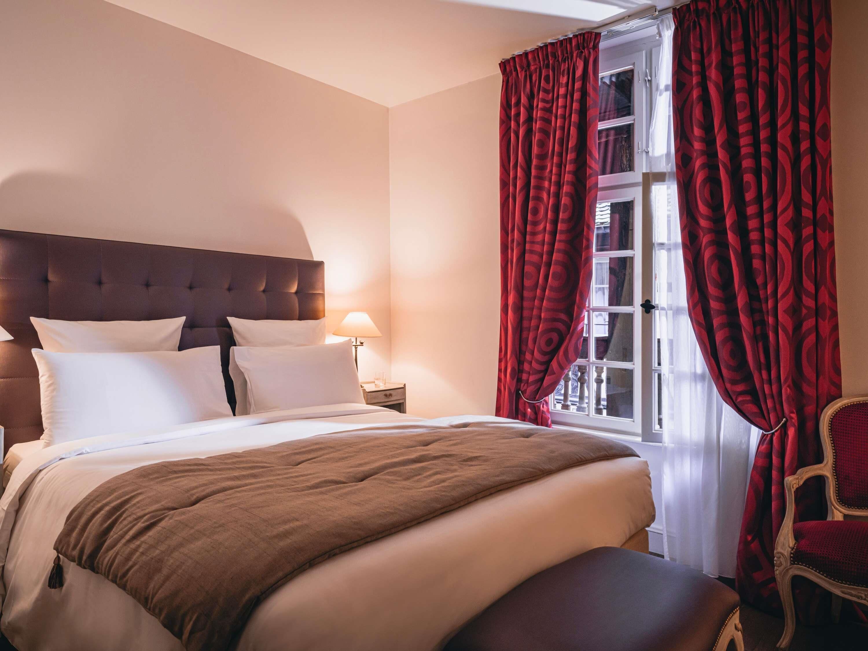 A hotel room with a large bed, a wooden headboard, a nightstand with a lamp, and red patterned curtains.
