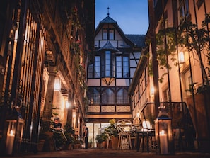 Una calle estrecha con edificios tradicionales de entramado de madera, mesas al aire libre y plantas en macetas.