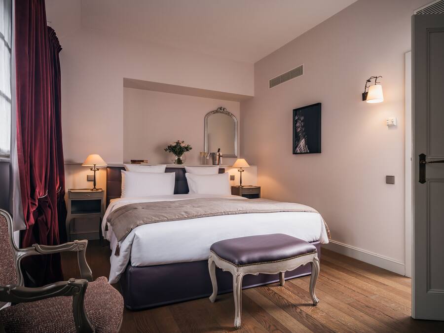 A bedroom with a large bed, bedside tables, a chair, a bench, a mirror, and a door.