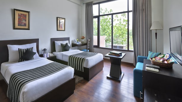 A hotel room with two beds, a desk, a chair, a window with curtains, and a view of greenery outside. - Regale Inn (New Delhi)