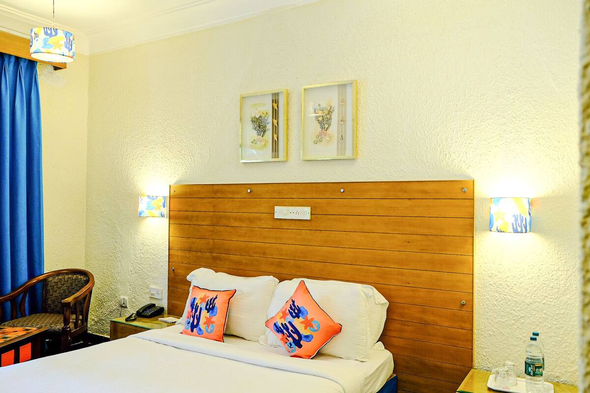 A hotel room with a wooden headboard, two framed floral pictures, a bed with white linens, two orange pillows, a wooden chair, a nightstand with a phone, and a table with a water bottle.