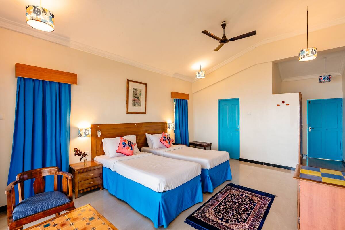 A hotel room with two beds, a wooden chair, a desk, a refrigerator, and a ceiling fan.