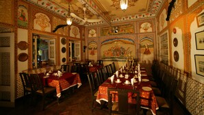 A traditional dining hall with ornate decorations, a long table set for a meal, and a ceiling fan. - Bissau Palace (Jaipur)