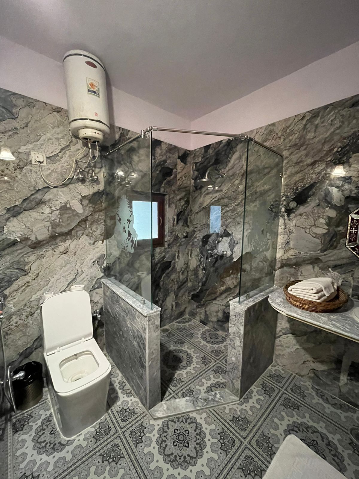 Un baño con pared de mármol, cabina de ducha de vidrio, inodoro y lavamanos con cesta para toallas.